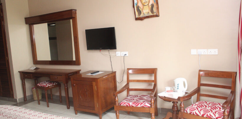 Image showing a chair, wall mounted TV, Electric Kettle, Mini-cupboard/desk and a wall hanging all in a room in the Mombasa Sports Club Accommodation Wing.